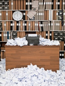 Photo of an office, desk in the middle overflowing with, and surrounded by, scrunched up paper. Typewriter on desk. Behind the desk is a bookcase full of books and folders and a clock