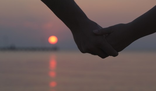 Close up of the hands of a romantic couple holding hands at sunset silhouetted against the colourful night sky with the orb of the sun visible below