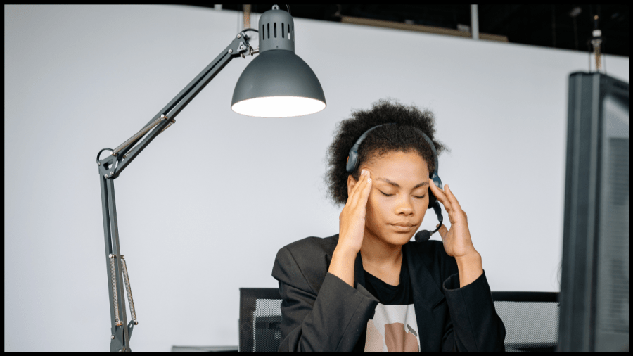 Photo of a young woman in front of a computer and wearing a headset, rubbing her temples.