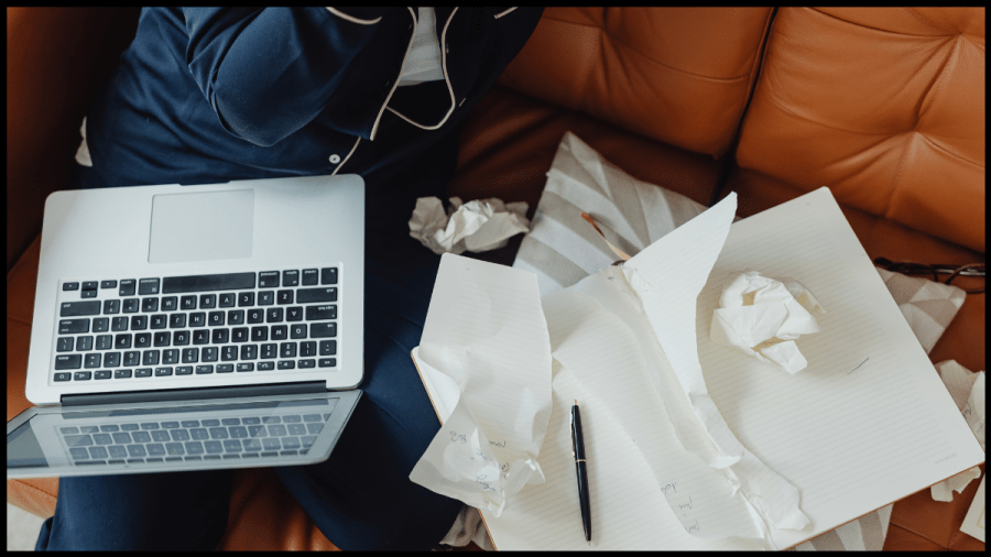 Image a laptop on a person's lap as they are sat on the sofa and papers strewn beside them.