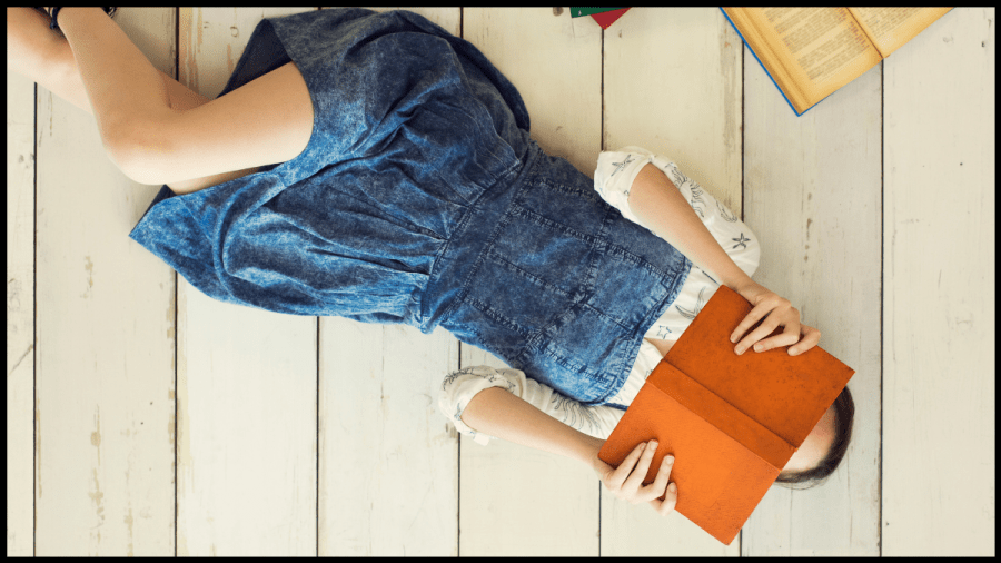 Photo of a young woman in a blue denim dress lying on a wooden floor holding up an orange covered book over her face