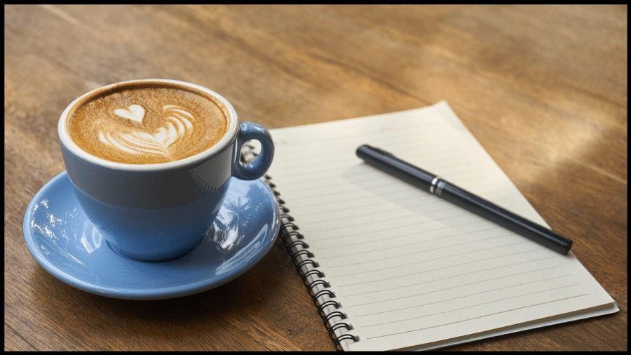Photo of a blue coffee cup and saucer beside a blank notepad with a pen sitting on a desk