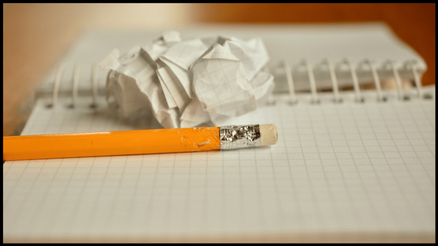 Close up photo of a yellow pencil with an erasure on top, lying on a blank notepad with a scrunched up ball of paper beside.