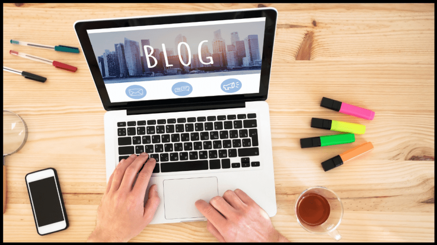 Photo of an open laptop on a desk with phone and highlights beside. A website is open with the word "blog" written across.