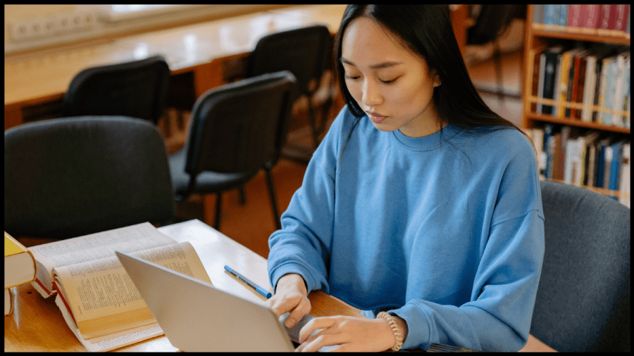Photo of a young woman in a blue sweatshirt, working on a laptop in a school setting.