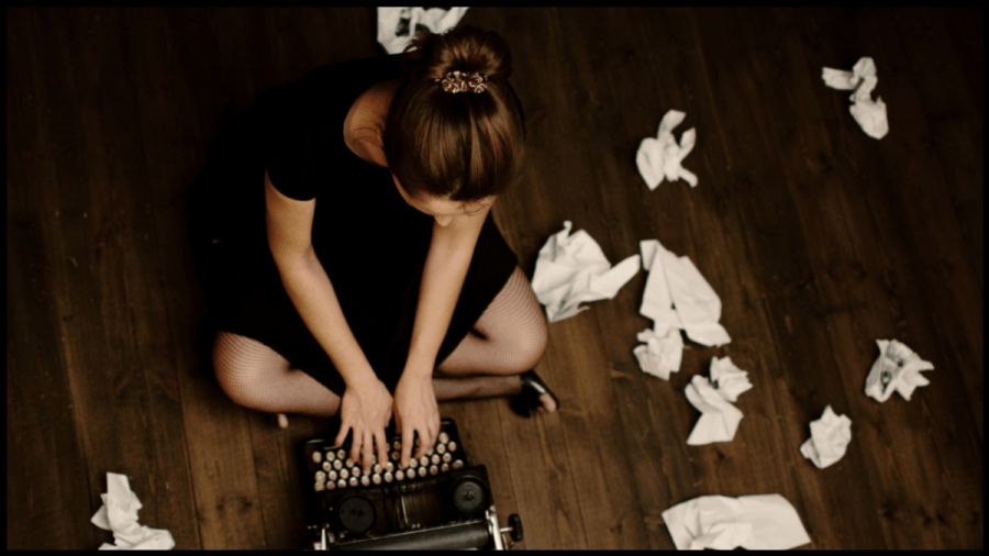 Photo of a young woman sitting cross legged on a wooden floor surrounded by balls of scrunched up paper while she types on a typewriter.