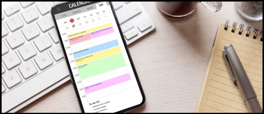 Close up photo of a phone, siting on a desk, with a calendar app open. There is also a laptop, pen and pad on the desk