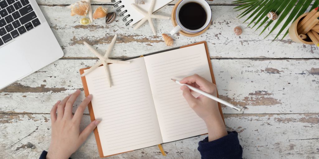 Image of a flatlay, a note book open on a wooden desk, laptop, coffee cup and hands holding a pencil ready to write