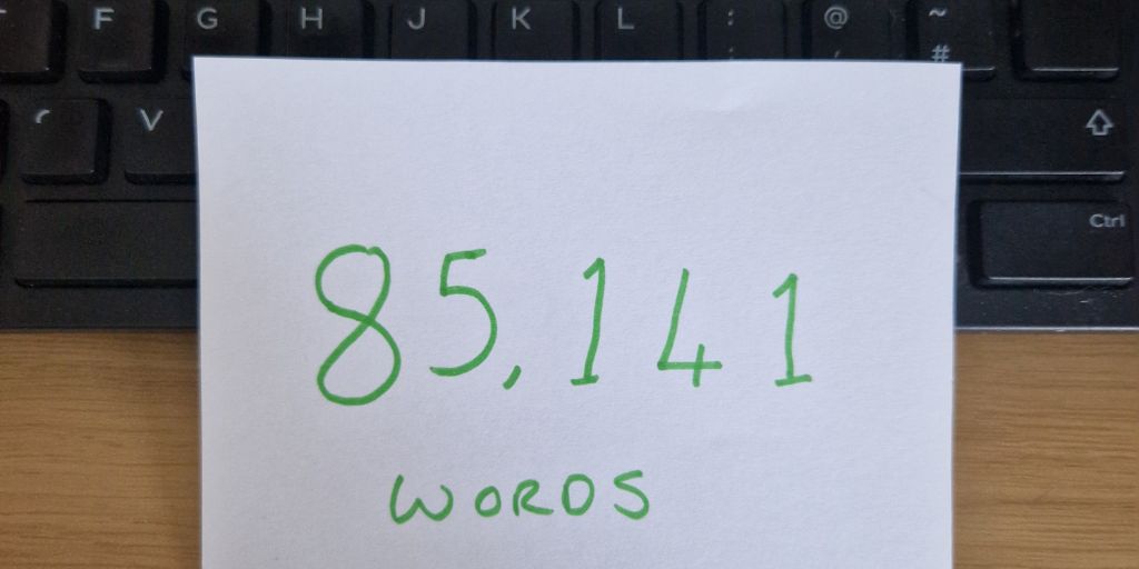 Photo of a slip of paper, resting up against a computer keyboard with 85,141 words written on it in green pen. 